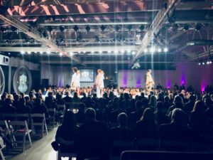 Fashion runway show with models walking under dramatic lighting and live audience, highlighting immersive sound and lighting design at a professional fashion event.