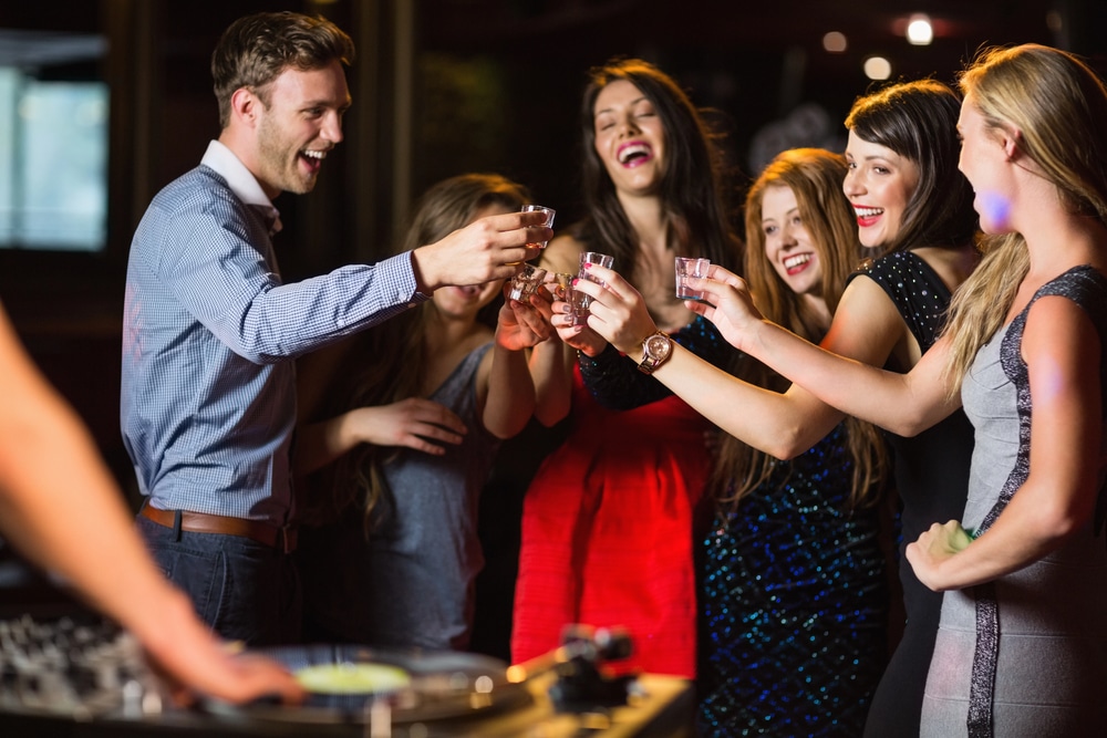 Group of friends toasting drinks by the DJ booth at a lively bar