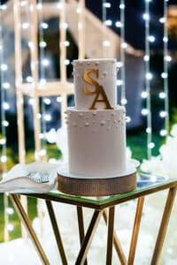 White wedding cake with gold monogram topper and lights behind